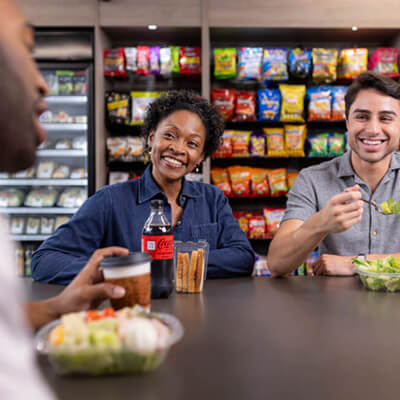People sitting at a table eating salads and snacks in front of shelves stocked with packaged foods.
