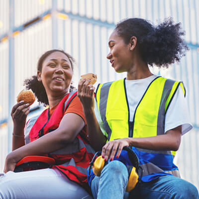 Two workers wearing safety vests sit outdoors near stacked shipping containers, holding sandwiches during a break, with a yellow hard hat placed on the ground.