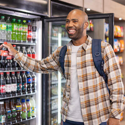 Person wearing a plaid shirt and backpack selecting a bottled soda from a refrigerated display stocked with various drinks and snacks in a modern self-service area.