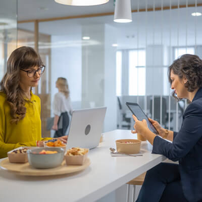 Two people sitting at a high table in an office, working with a laptop and tablet beside bowls of snacks.