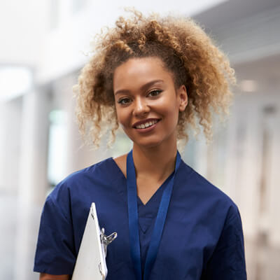Healthcare worker in navy scrubs with a lanyard holding a clipboard, standing in a bright hospital hallway