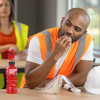 Person in an orange safety vest sitting at a table with a sandwich, a white hard hat, and a red sports drink bottle in a worksite break area.