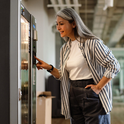 Person wearing a striped shirt and dark pants selecting an item from a vending machine in a modern indoor setting.