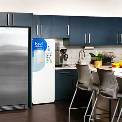 Modern office kitchen with dark cabinets, a stainless steel refrigerator, and a Bevi water dispenser next to a counter with chairs and a potted plant.