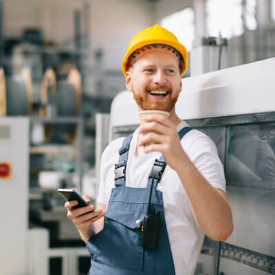 Factory worker wearing a yellow hard hat and blue overalls holds a coffee cup and a smartphone while standing near industrial equipment.