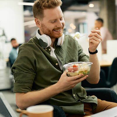 Man sitting in an office eating a salad from a clear bowl while wearing headphones around his neck.