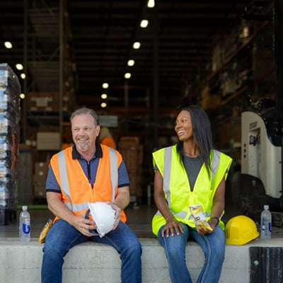 Two warehouse workers in safety vests sitting on a loading dock, taking a break with water bottles and snacks near stacks of packaged goods.
