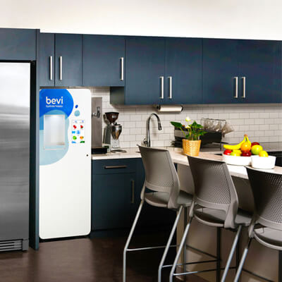 Modern office kitchen with dark blue cabinets, a Bevi water dispenser, stainless steel fridge, countertop with fruit bowl, and gray bar stools.