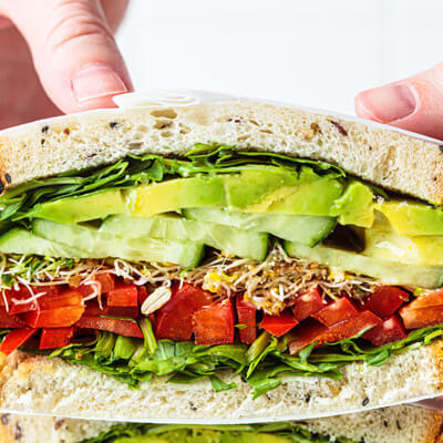 Close-up of a fresh vegetable sandwich layered with avocado, cucumber, sprouts, red peppers, and greens on multigrain bread.