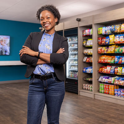Person standing with arms crossed in front of a workplace snack market with shelves of snacks and a self‑checkout kiosk.