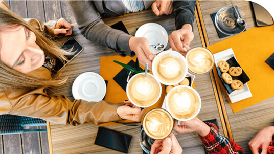 Top view of several people clinking cups of cappuccino over a wooden table with saucers, cookies, and smartphones.