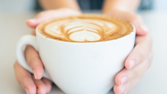 Hands holding a white cup of coffee with heart-shaped latte art on a light surface.