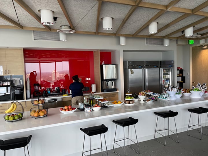 Modern office pantry with a long white counter, black stools, baskets of fresh fruit, bowls of snacks, coffee machines, and refrigerators against a red backsplash.