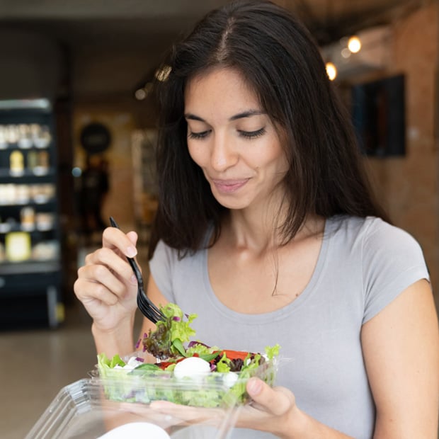 Woman holding a plastic container of salad while eating with a black fork indoors.
