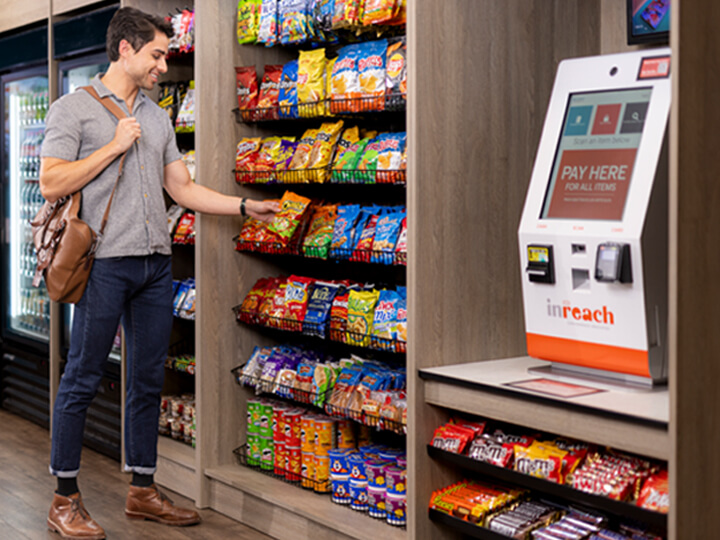 Person selecting a bag of chips from a snack display next to a self-service payment kiosk in a micro market.