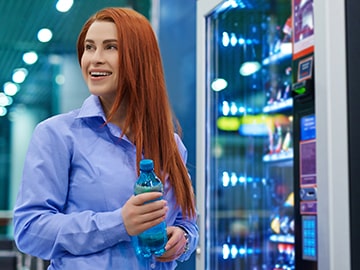 Person holding a bottled drink while standing beside a brightly lit vending machine.