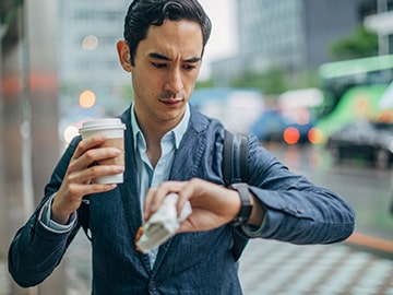 Person standing on a city sidewalk holding a coffee and checking the time while carrying a backpack.