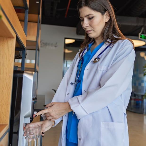 A healthcare worker in scrubs and a white coat dispenses water into a glass from a dispenser in a modern office setting.