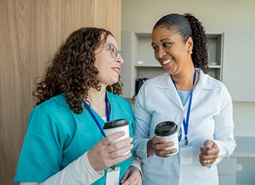 Two healthcare workers in uniforms holding coffee cups while talking in a break room.