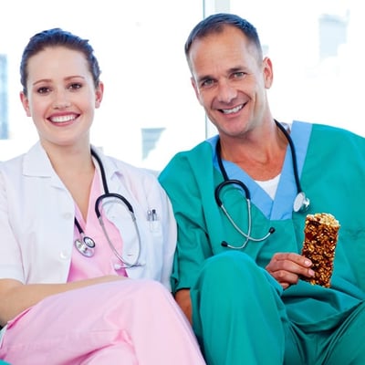 Two healthcare workers in scrubs sitting together, one holding a granola bar.
