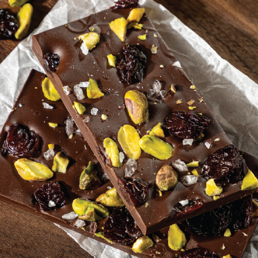 Dark chocolate bark topped with pistachios, dried cherries, and coarse sea salt, arranged on parchment paper over a wooden surface.