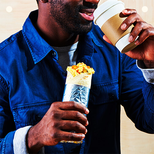 A man enjoys a cup of coffee while holding a sandwich