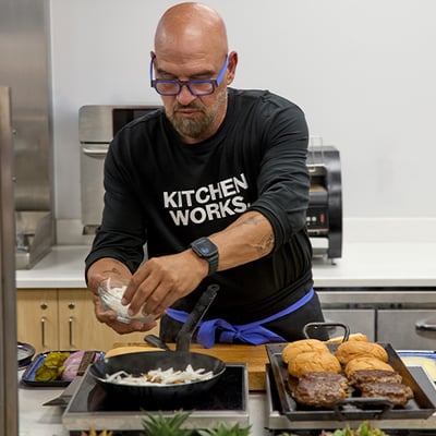 Person wearing “Kitchen Works” shirt adds sliced onions to skillet; burger patties and buns rest on nearby tray.