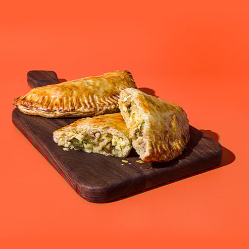 Two golden-brown empanadas on a dark wooden board, one halved to show a savory filling of vegetables and meat, set against an orange background.
