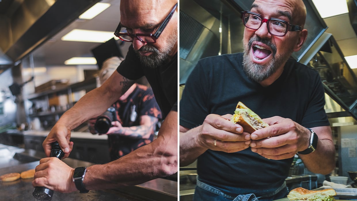 A man with glasses and a beard holds a sandwich, smiling as he prepares to take a bite.