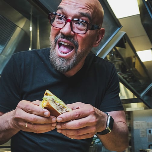 A man with glasses and a beard holds a sandwich, smiling as he prepares to take a bite.
