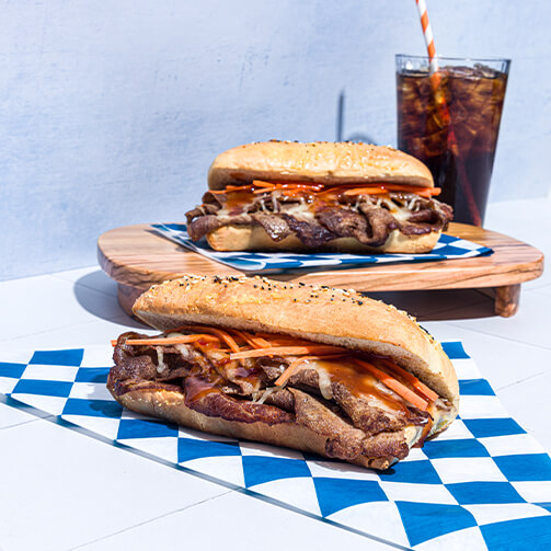 Two toasted sandwiches filled with sliced beef, shredded cheese, and vegetables on blue checkered paper, served with a glass of cola and a striped straw.