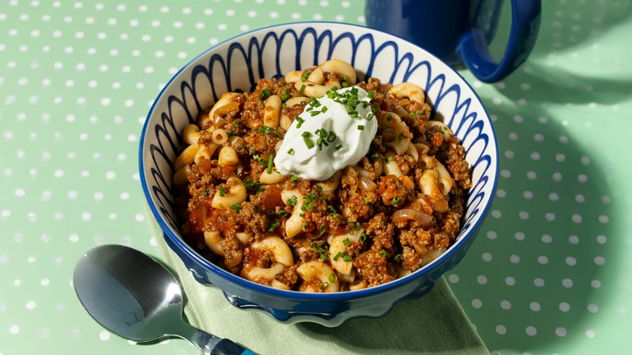 Bowl of macaroni pasta mixed with ground meat and tomato sauce, garnished with chopped chives and a dollop of sour cream, set on a green polka-dotted surface with a spoon and napkin nearby. A blue mug sits in the background.