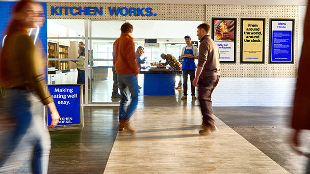 A group of people walking through a busy kitchen, engaged in various cooking and food preparation activities.