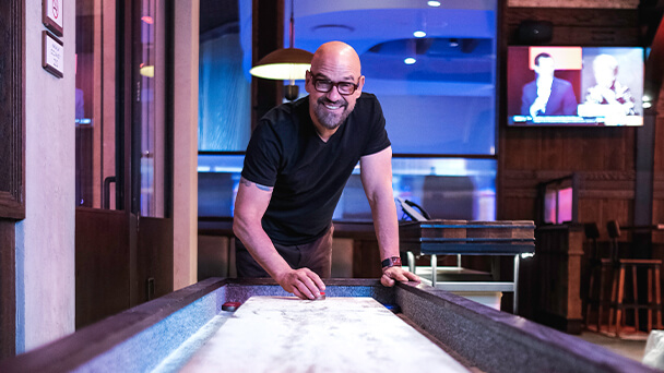 A man wearing glasses plays shuffleboard in a lively restaurant setting.