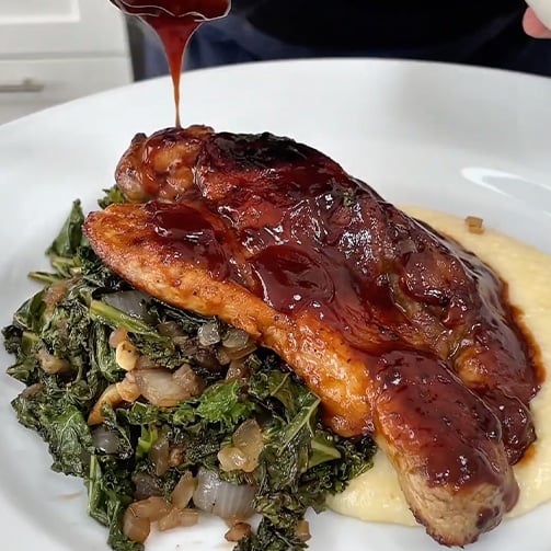 Close-up of a plate with slow-roasted pork butt steak with cherry BBQ sauce, creamy cheddar grits and balsamic collard greens.  