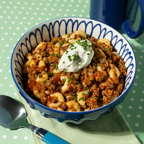 A bowl of chili topped with sour cream accompanied by a spoon set against a neutral background