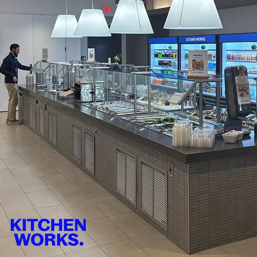 Person serving themselves at a salad bar in a modern cafeteria, with "KITCHEN WORKS." text in the corner.