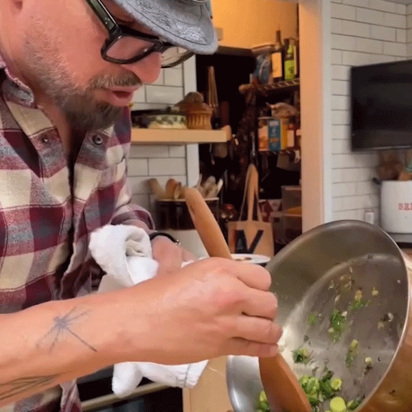 A man wearing a hat and glasses skillfully cooks food in a pan over a stove