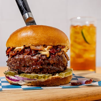 Close-up cheeseburger topped with crispy bacon and sauce, served on a bun with a drink in the background.