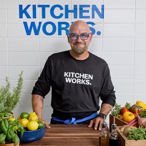 Chef Michael Symon standing behind a kitchen counter with fresh produce, wearing a “KITCHEN WORKS.” shirt.