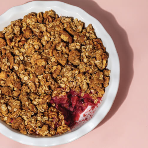 White dish filled with golden-brown oat and nut crumble, partially revealing a layer of vibrant red fruit filling, set against a soft pink background.