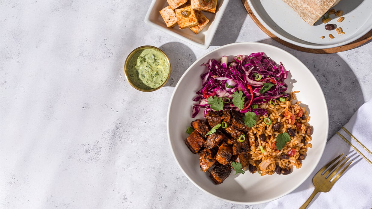 Bowl with rice, beans, red cabbage slaw, tofu cubes; green sauce and a wrap on plates nearby.
