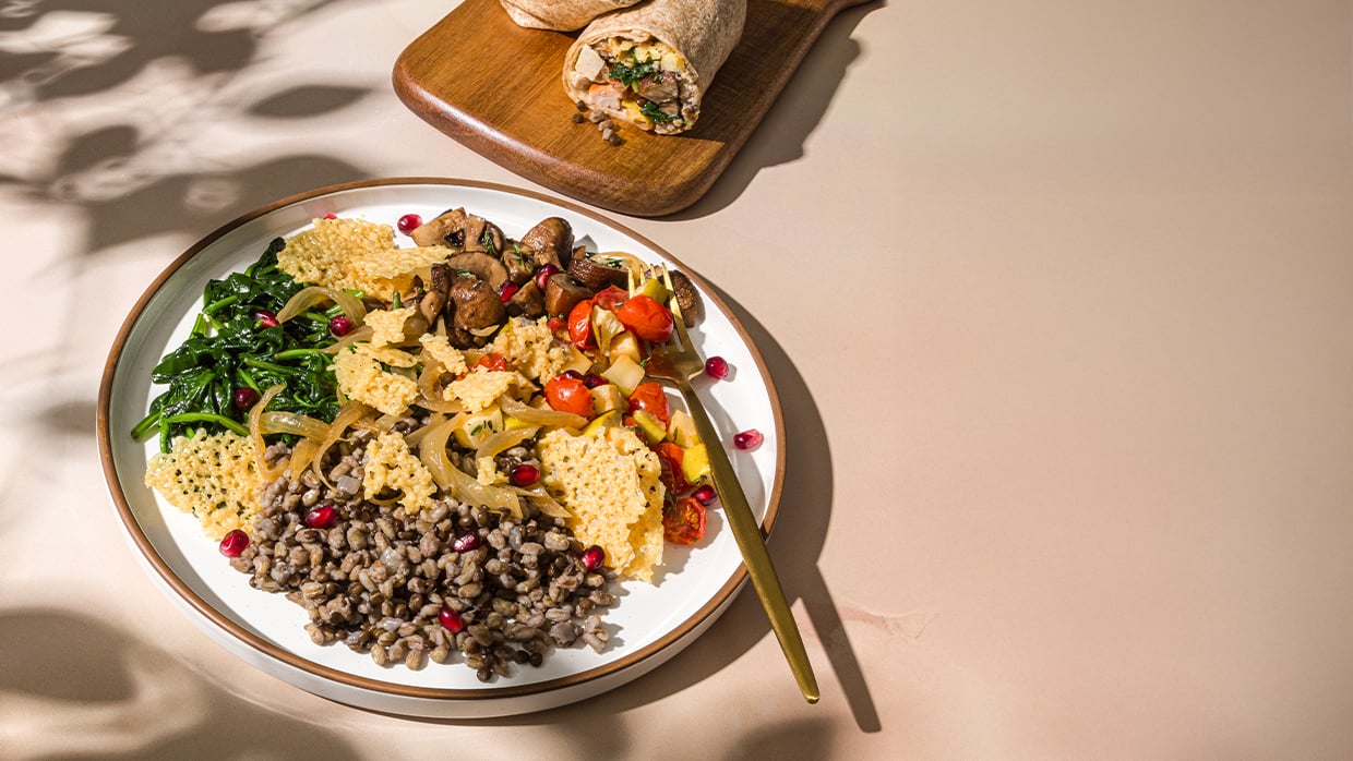 Plate of veggies, grains, cheese crisps with fork, beside wooden board holding a sliced wrap.