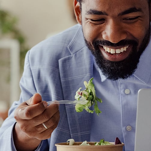 Person in a light blue suit jacket eating salad with a fork from a bowl, with part of a laptop visible on the side.