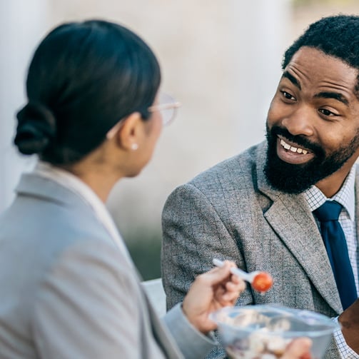 Two individuals in formal attire sitting and conversing; one holds a fork with food and a bowl of salad.