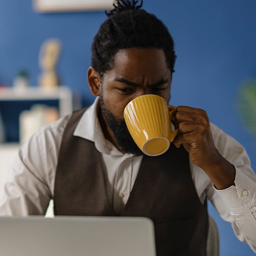 Person in a white shirt and dark vest holding a yellow mug while sitting in front of a laptop, with a blue background and shelves behind.