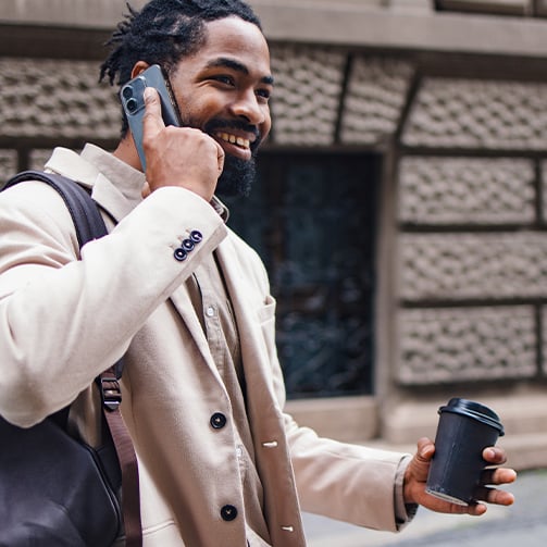 Person in a light-colored coat holding a smartphone to their ear and a black coffee cup, with a black shoulder bag in an urban setting.