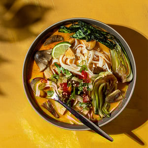 Bowl of noodle soup with vegetables, lime, and chili peppers on a yellow background.