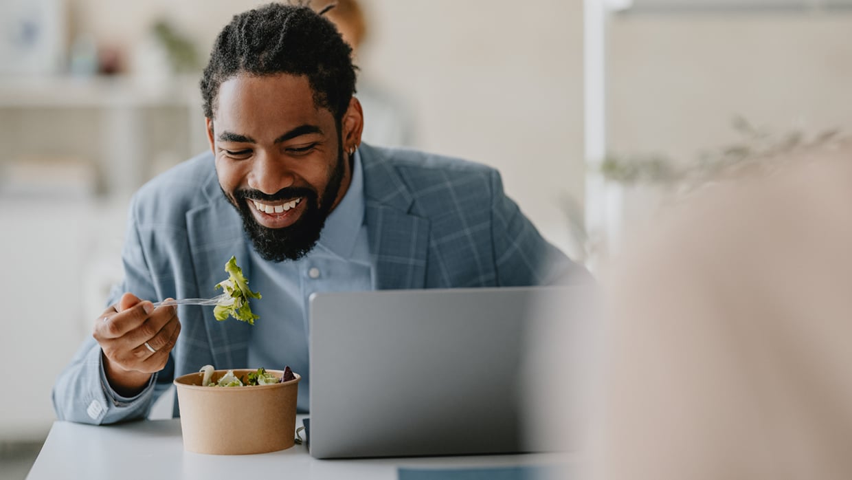 Person wearing a light blue suit jacket and a light blue shirt, seated at a table with a laptop, holding a fork with a piece of lettuce while eating from a brown paper bowl filled with salad. Background suggests an indoor setting, possibly an office or cafeteria