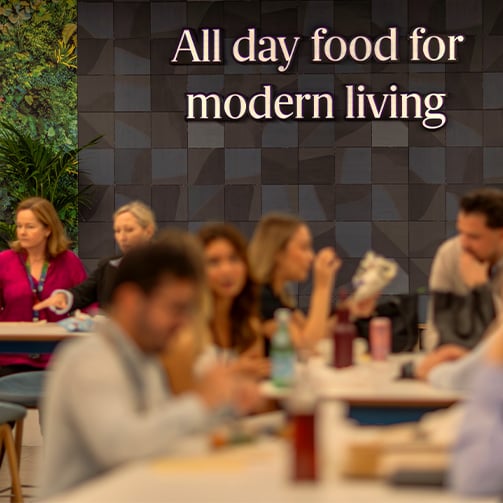 People sit in a cafeteria with a green plant wall and a sign reading “All day food for modern living.”